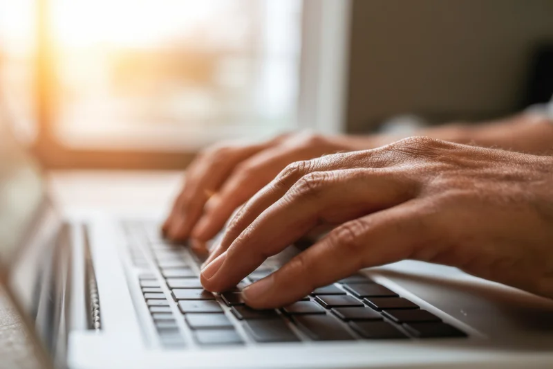 Hands typing on a laptop keyboard by a sunlit window