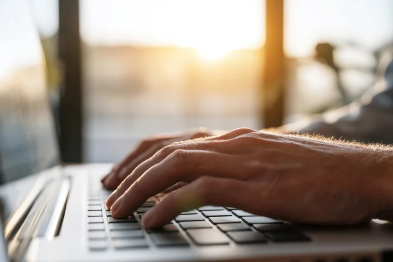 Hands typing on a laptop keyboard in warm sunlight at evening