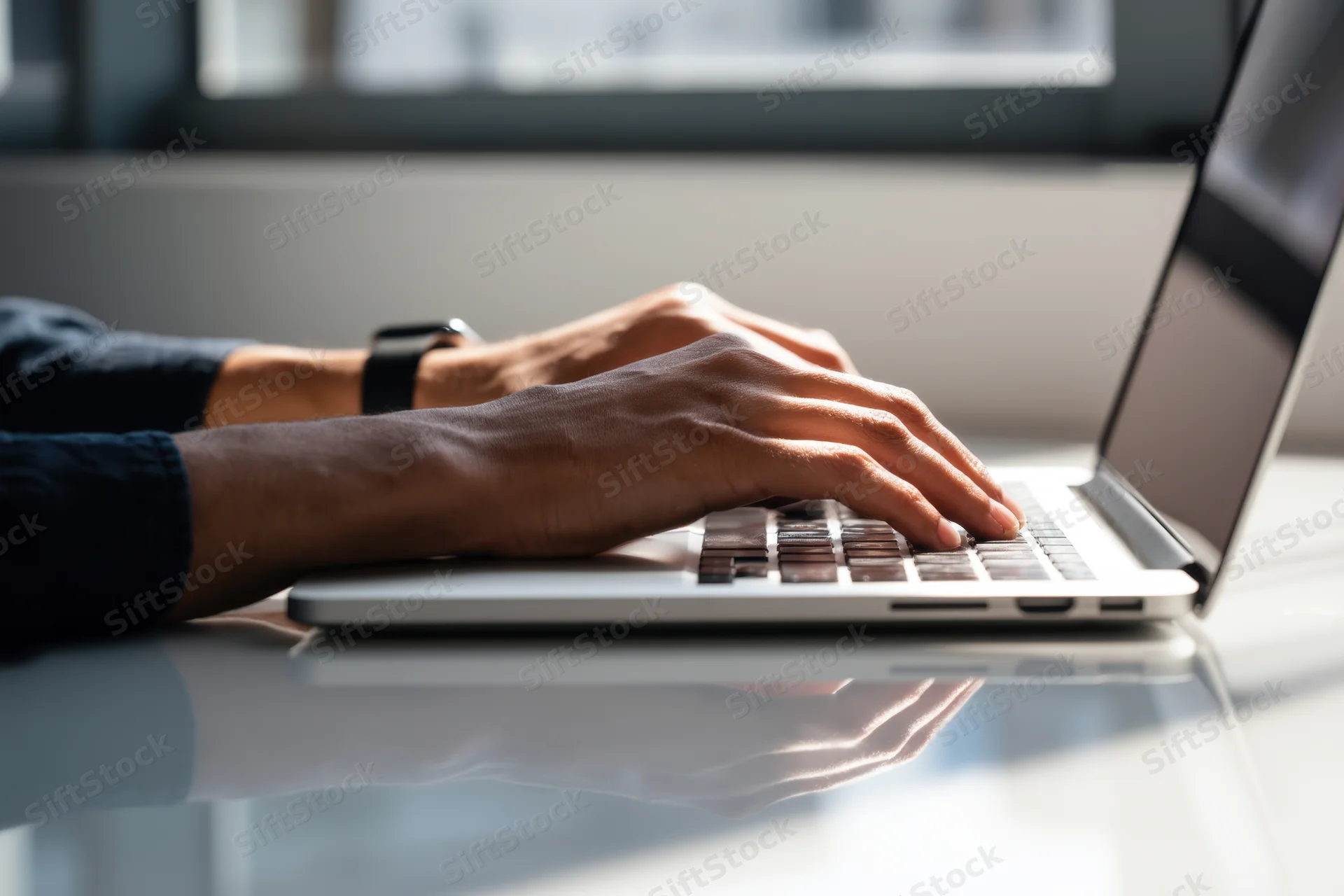 Hands typing on a silver laptop at a glass desk in a bright office