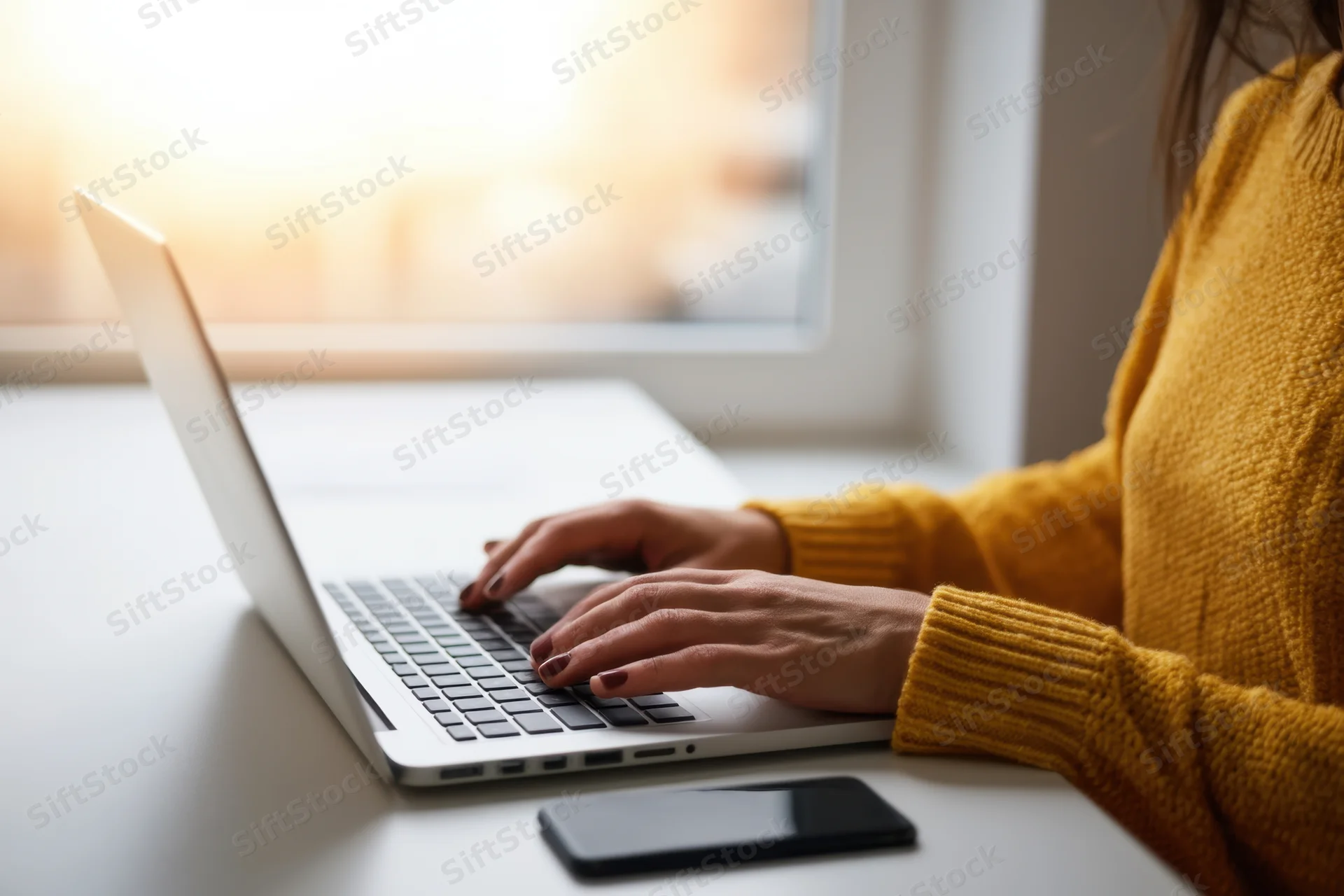 Woman typing on a silver laptop at a clean desk by a window