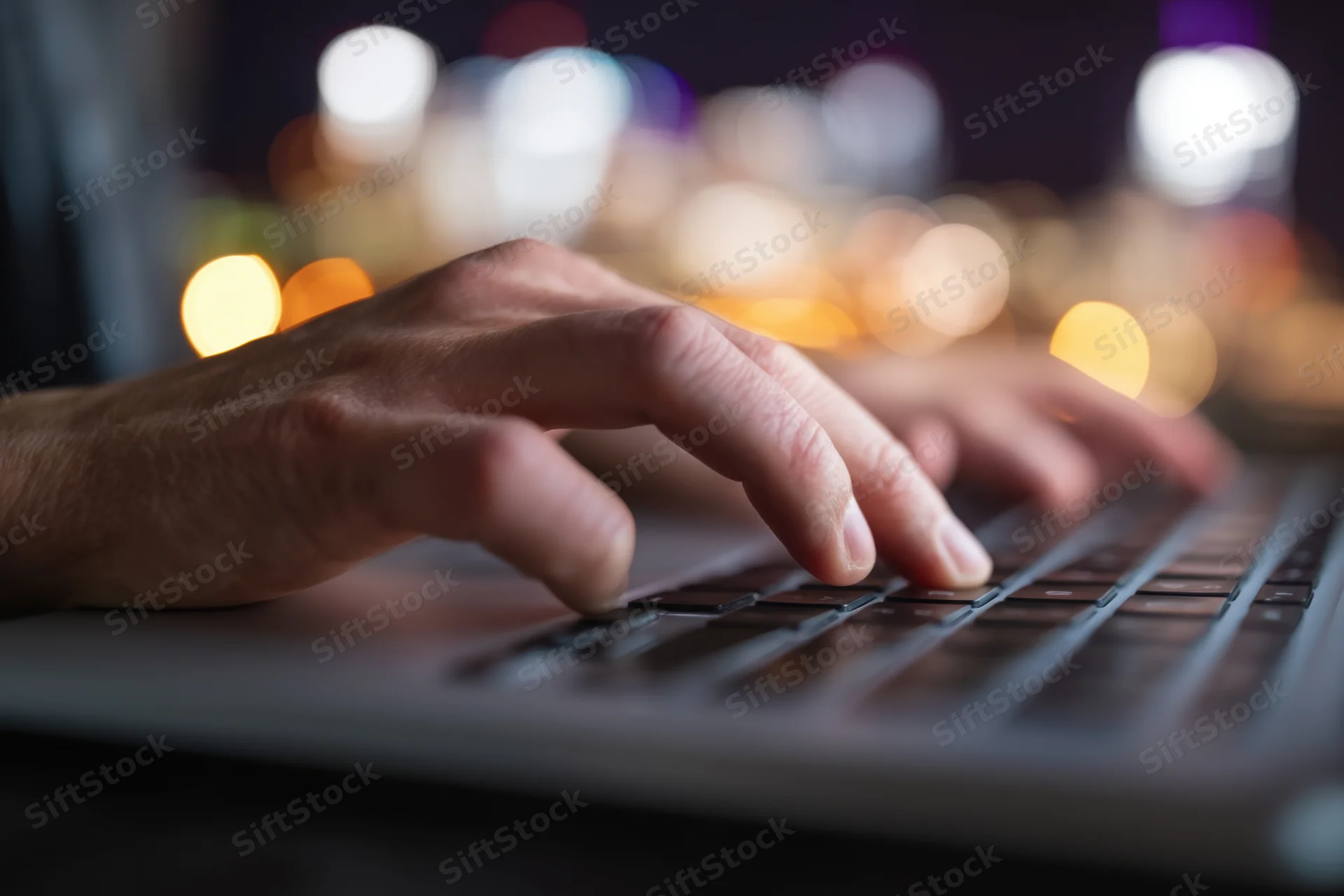 Hands typing on a laptop keyboard at night with city lights blurred in the background