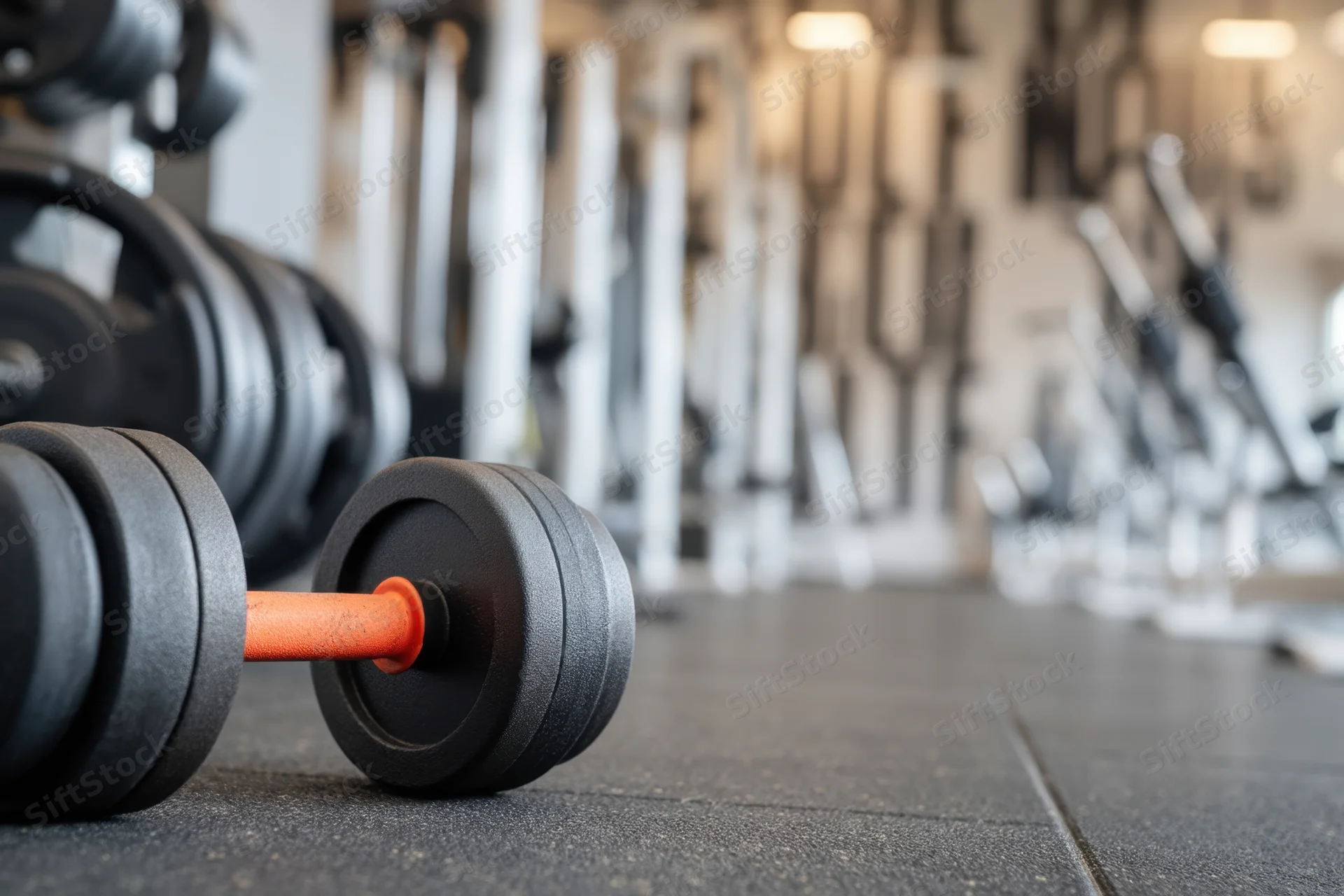 Close-up of a dumbbell with black rubber plates and orange handle resting on a gym floor, surrounded by exercise equipment in the background