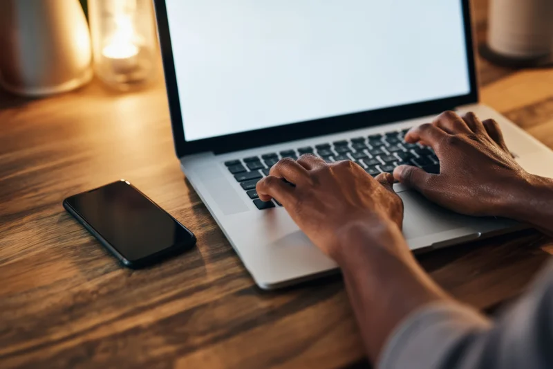 Hands typing on a silver laptop on a wooden desk with a smartphone beside it