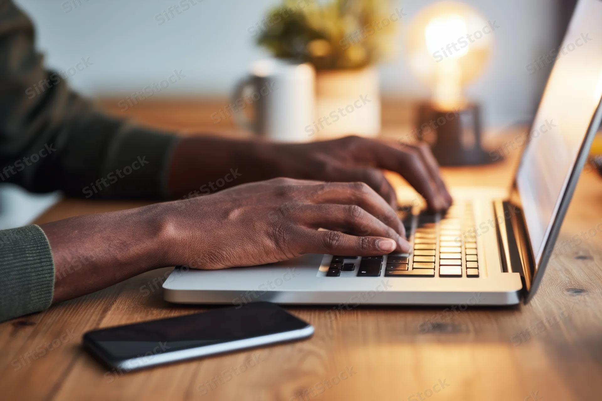 Close-up of hands typing on a silver laptop on a wooden desk with a smartphone and lamp nearby