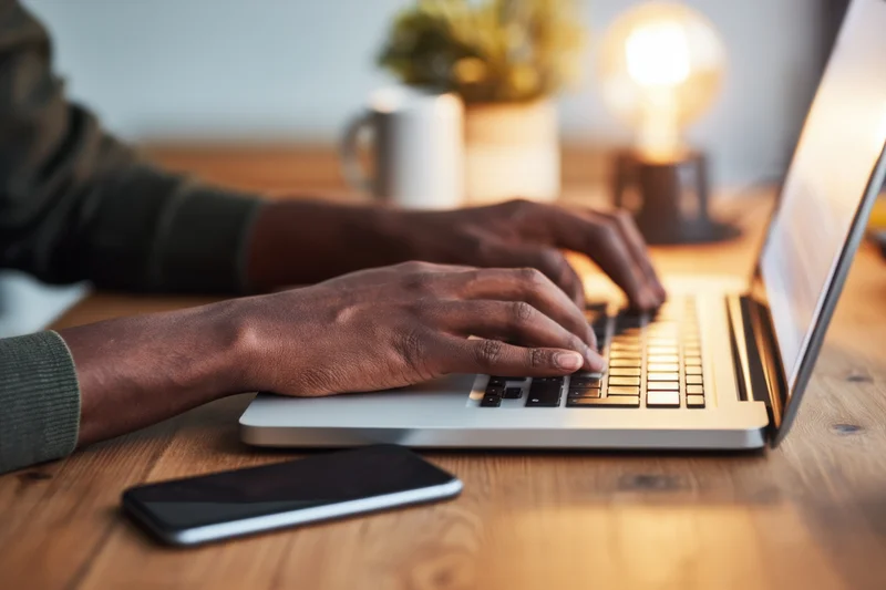 Close-up of hands typing on a silver laptop on a wooden desk with a smartphone and lamp nearby