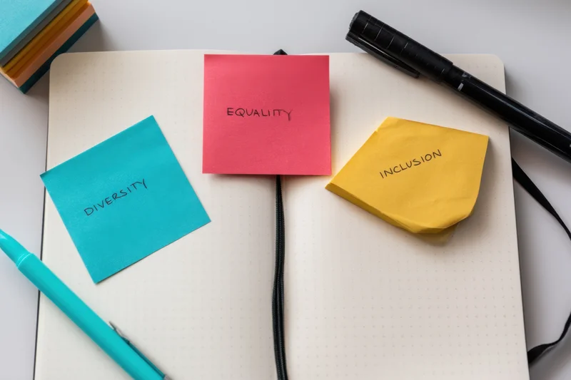 Three colorful sticky notes labeled diversity, equality, and inclusion placed on an open dotted notebook with pens on a white desk