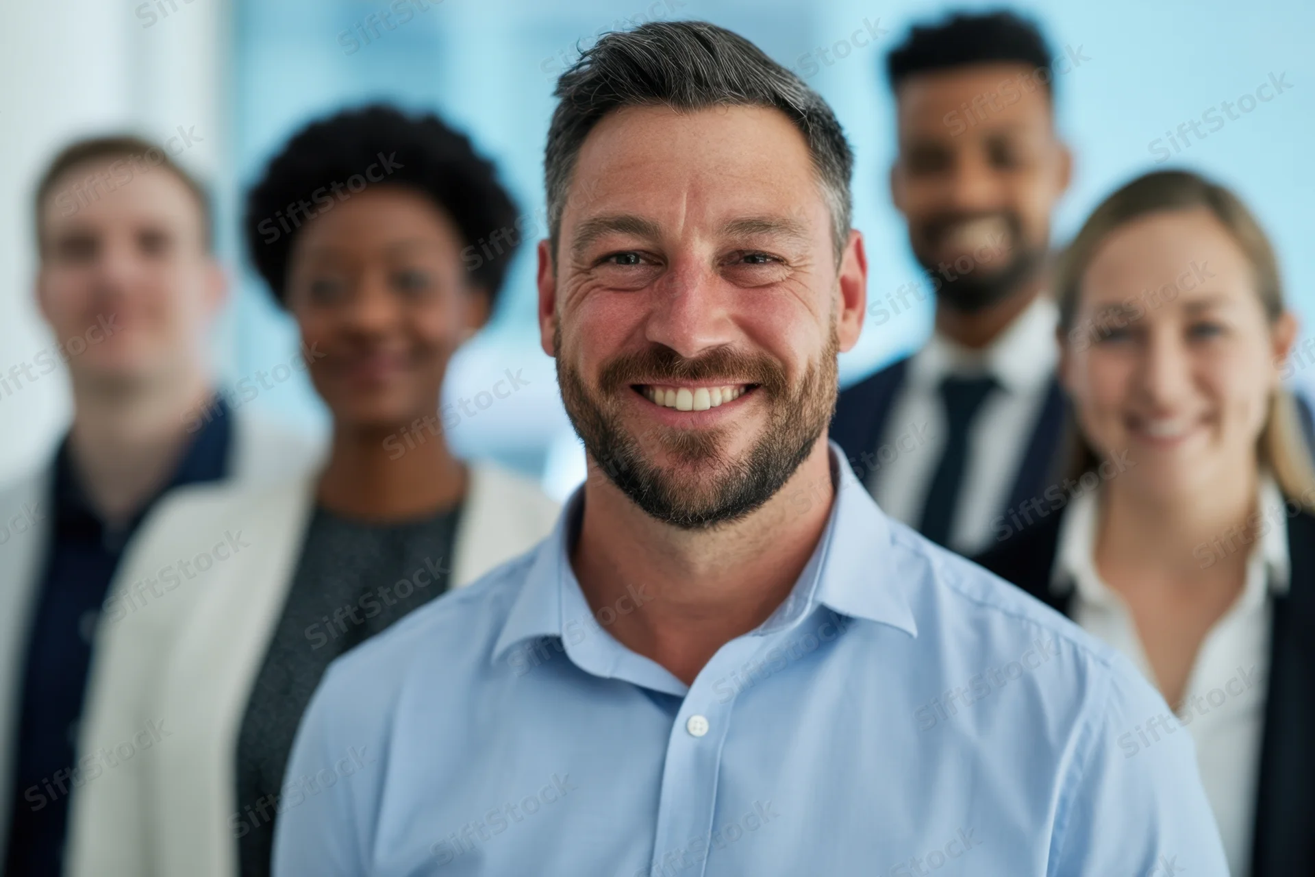 Smiling man in light blue shirt leading a diverse team of professionals in a bright office setting