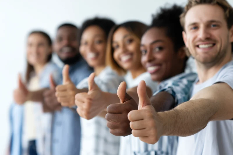Diverse group of people giving thumbs up in a row, smiling, in a bright indoor setting