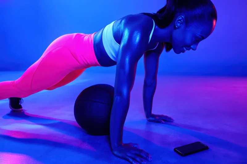 Woman doing pushup in plank position on floor with medicine ball and smartphone nearby