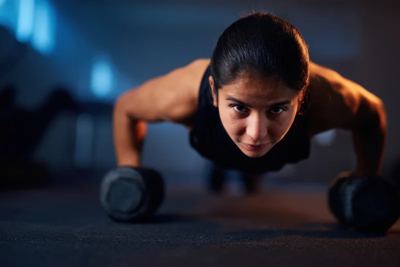 Woman performing weighted pushup with dumbbells in dimly lit gym