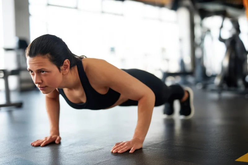 Woman doing pushup on gym floor in black sports bra and leggings