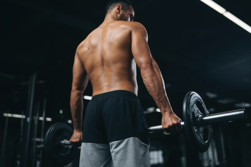 Muscular man lifting barbell in gym, back view, focused expression, wearing black and gray shorts