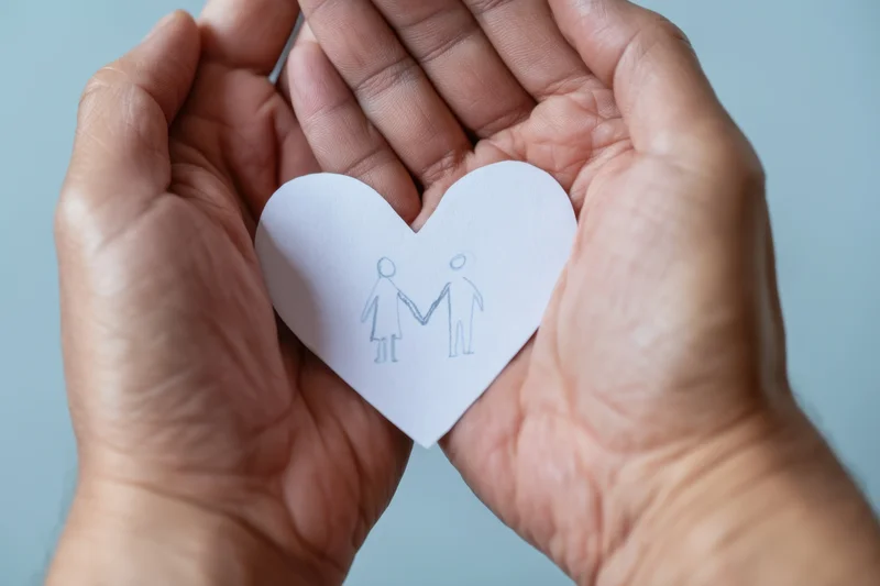 Hands cradling a paper heart with a couple drawing against a blue background