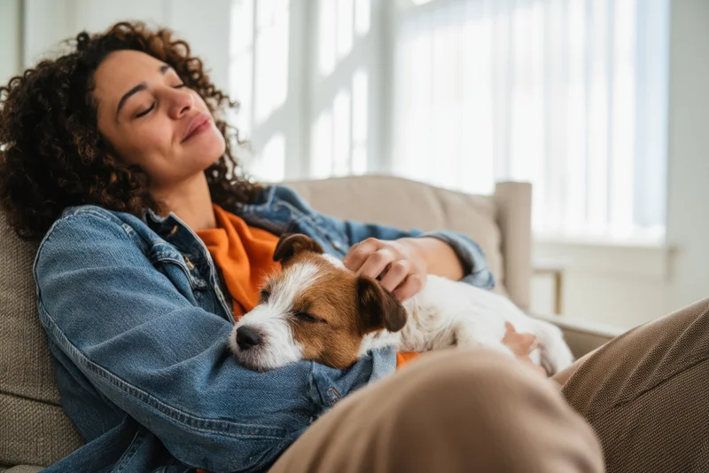 Woman resting with a sleeping dog on a couch in a sunlit living room