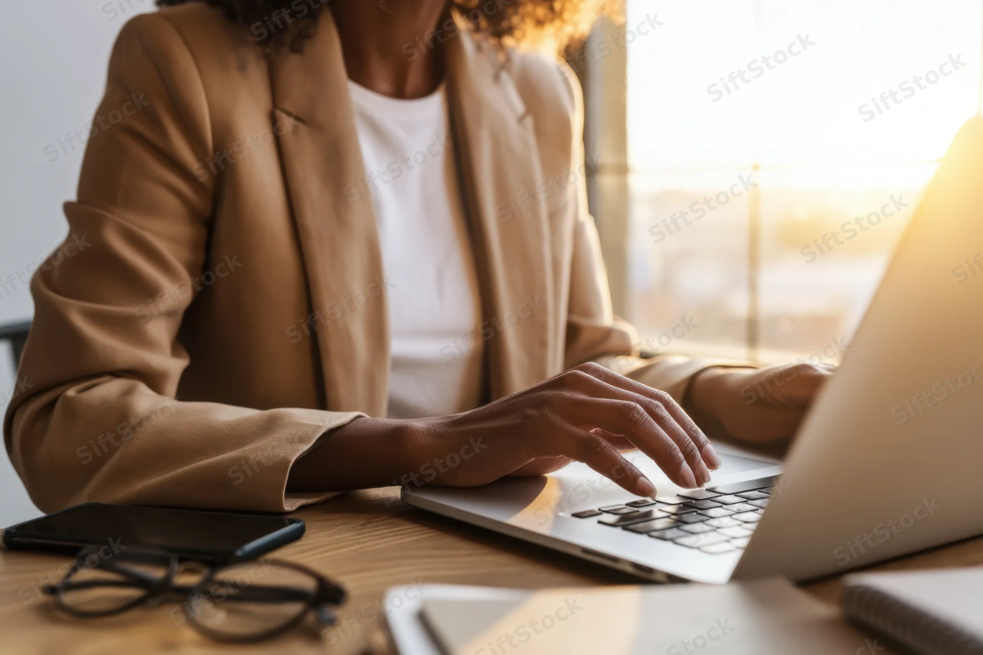 Woman in tan blazer typing on laptop at wooden desk by window