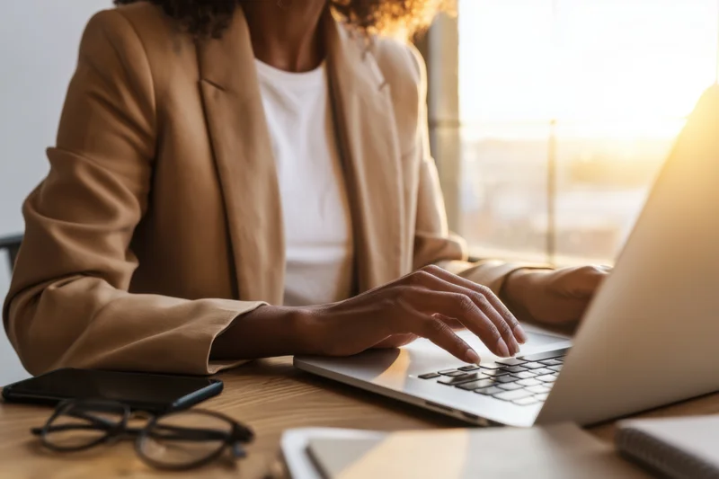 Woman in tan blazer typing on laptop at wooden desk by window