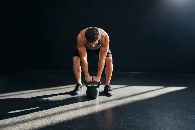 Man bending to grip kettlebell on dark gym floor with sunlight stripes