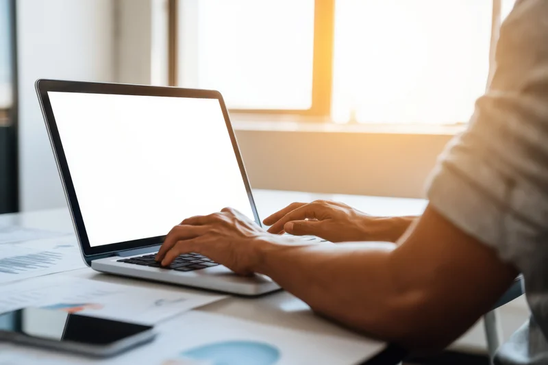 Person typing on laptop at bright desk with documents and blank screen