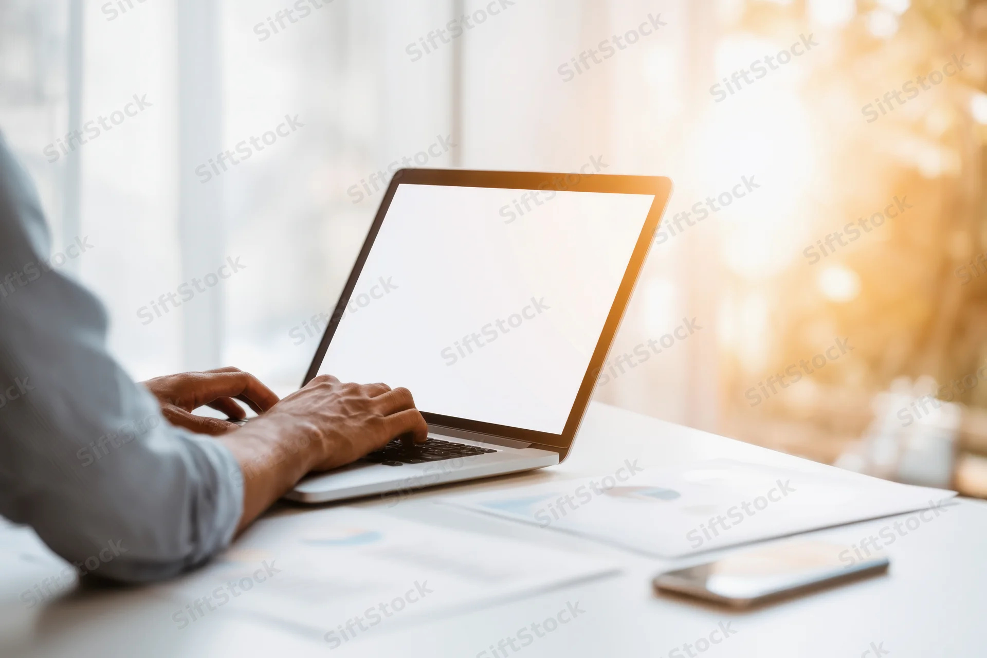 Man typing on laptop at bright desk with documents and smartphone nearby
