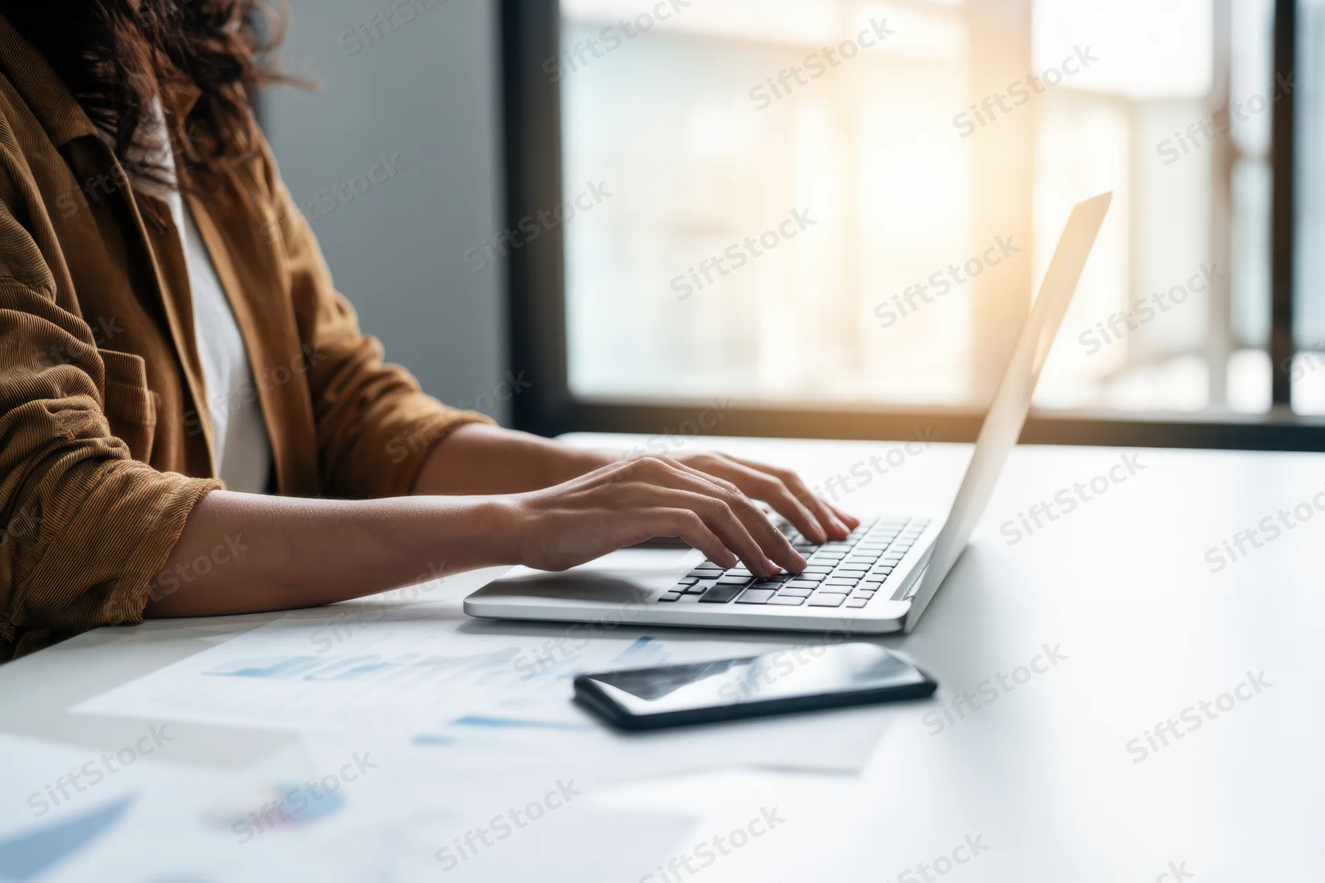 Woman typing on laptop at bright desk with documents and smartphone nearby