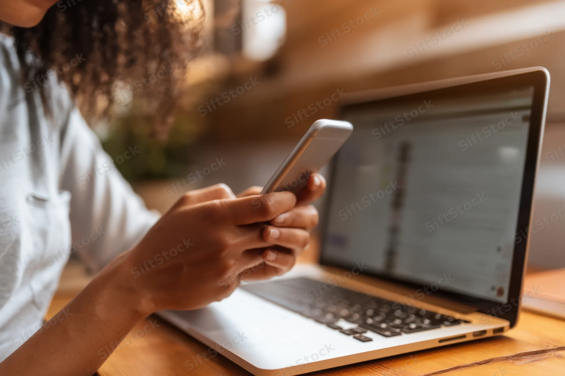 Woman holding smartphone while working on laptop at wooden desk