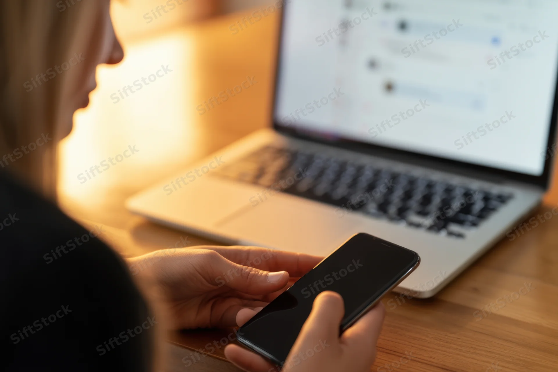 Woman holding smartphone while using laptop at wooden desk