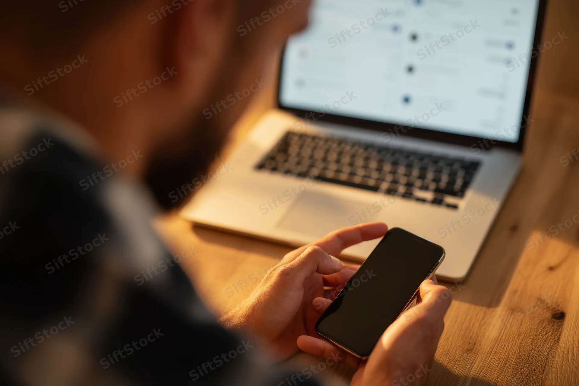 Man holding smartphone while using laptop at wooden desk