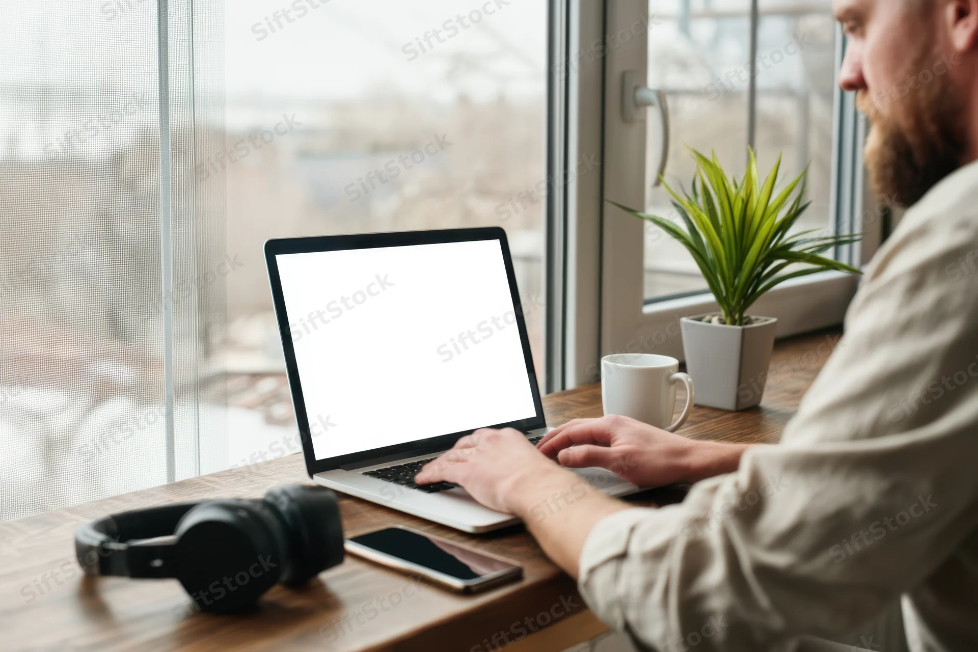 Man typing on laptop at wooden desk by large window with headphones and plant nearby