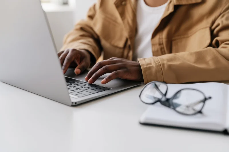 Person typing on a silver laptop at a white desk with notebook and glasses nearby