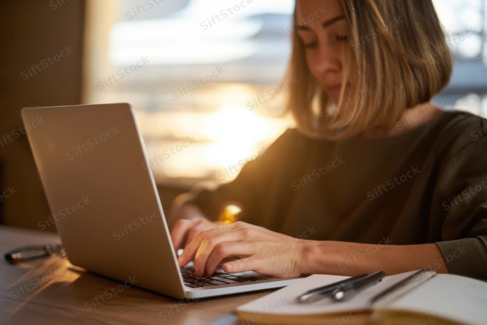 Woman typing on laptop at wooden desk in home office during golden hour