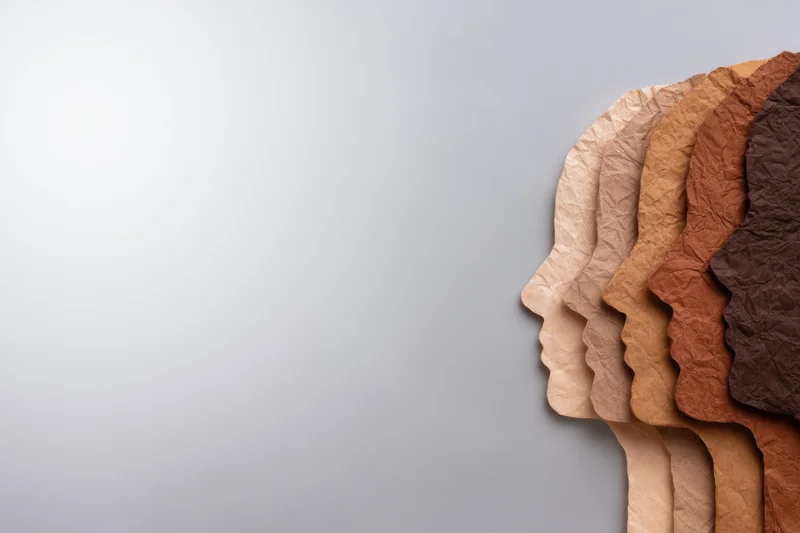 Row of crumpled paper head silhouettes in various skin tones arranged side by side on a gray background