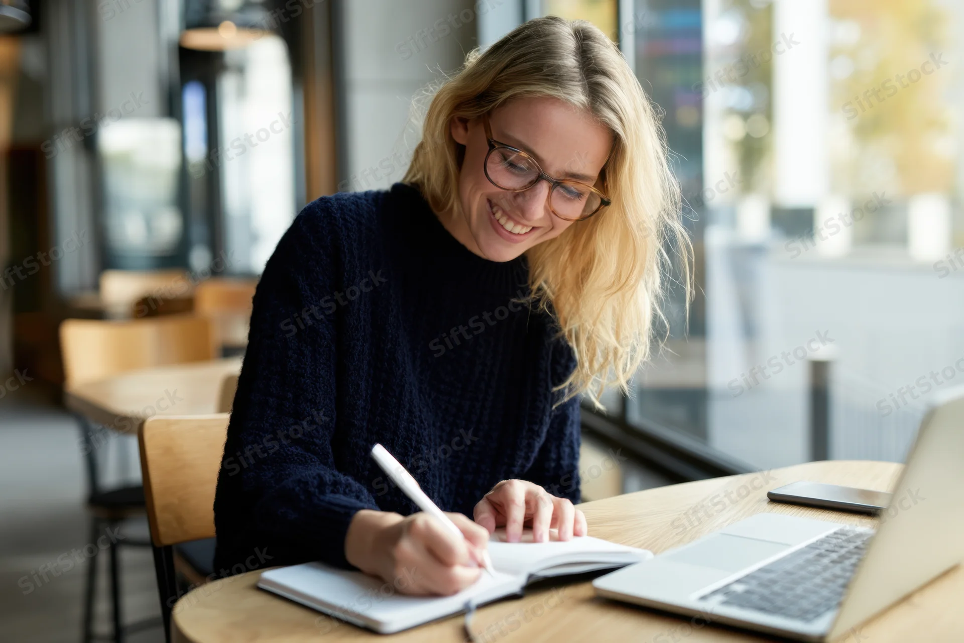 Smiling blonde woman writing in notebook at cafe table with laptop