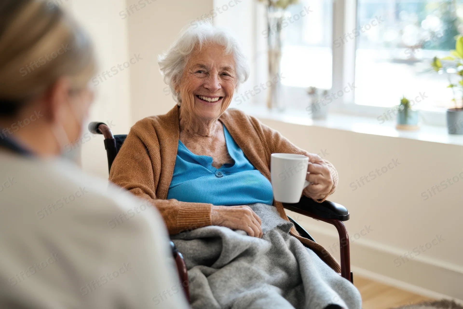 Elderly woman in wheelchair smiling while holding mug during conversation with caregiver indoors