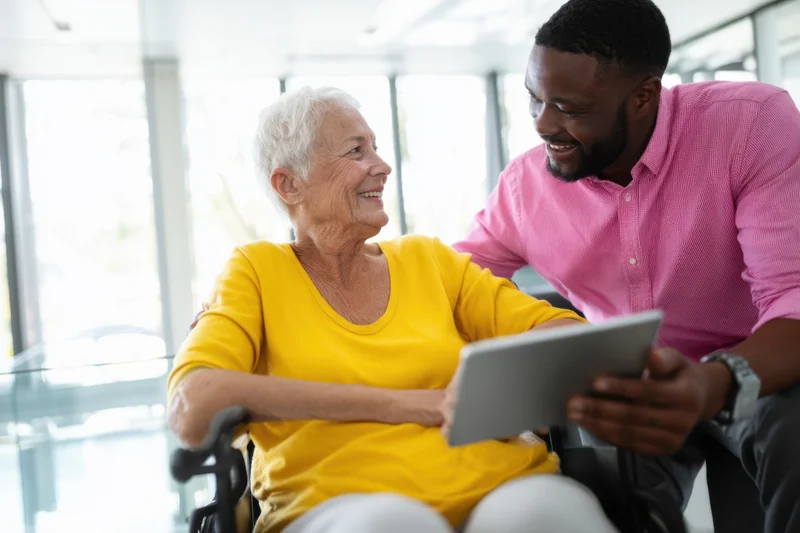 Elderly woman in wheelchair smiling while sharing tablet with caregiver in bright indoor setting