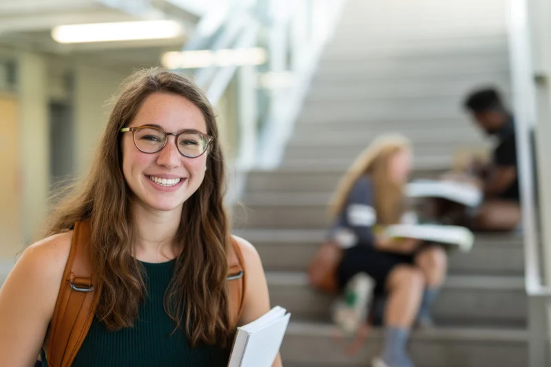 Smiling female student with glasses and backpack holding notebook in university hallway with peers on stairs in background