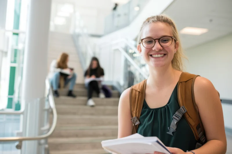 Smiling female student holding notebook in university hallway with peers on stairs in background