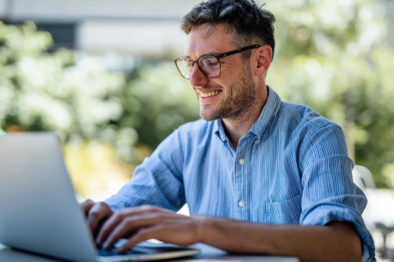 Man smiling while typing on laptop outdoors in casual setting