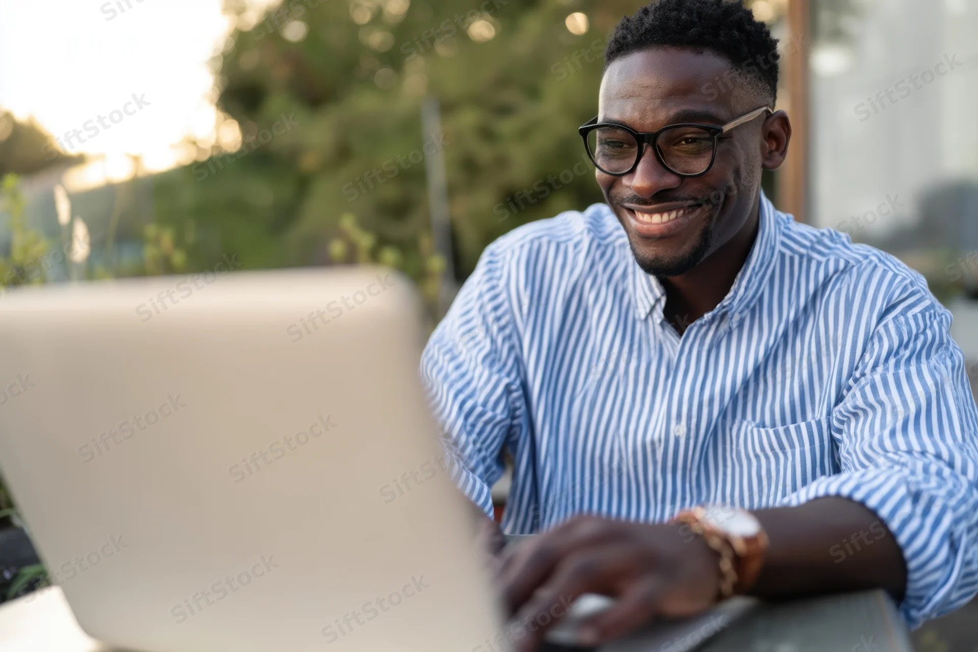 Smiling man typing on laptop outdoors in a green urban setting