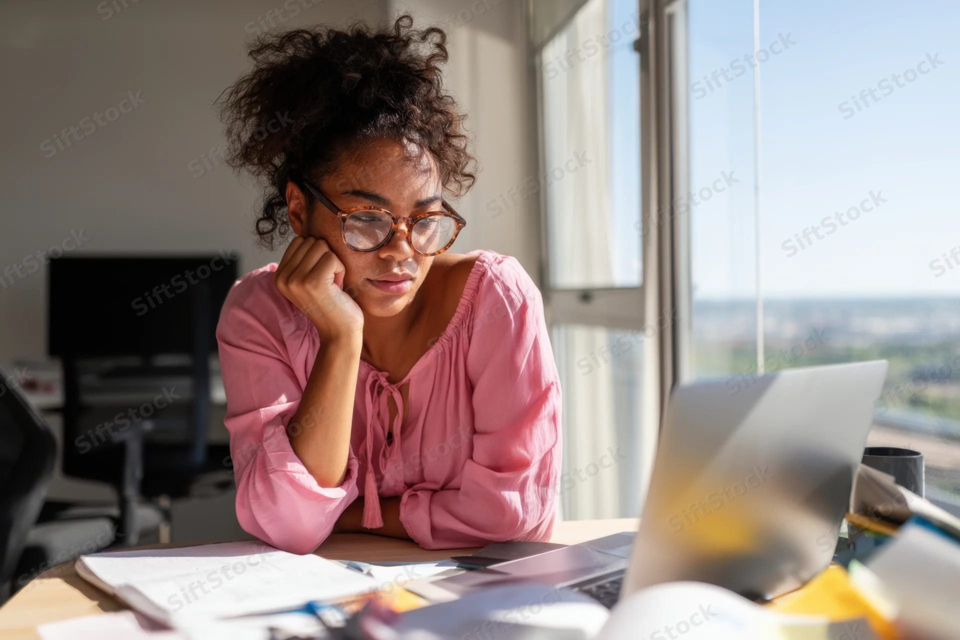 Woman in pink blouse concentrating at cluttered desk by window in office