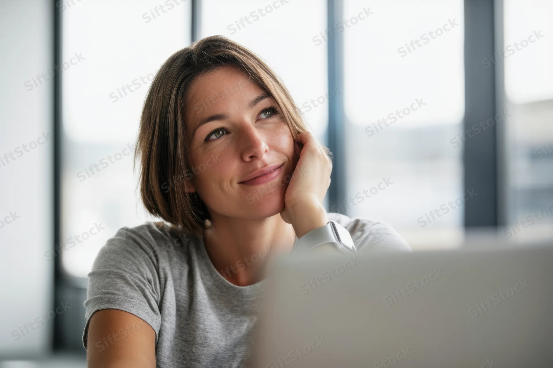 Young woman smiling thoughtfully at desk in front of laptop near window