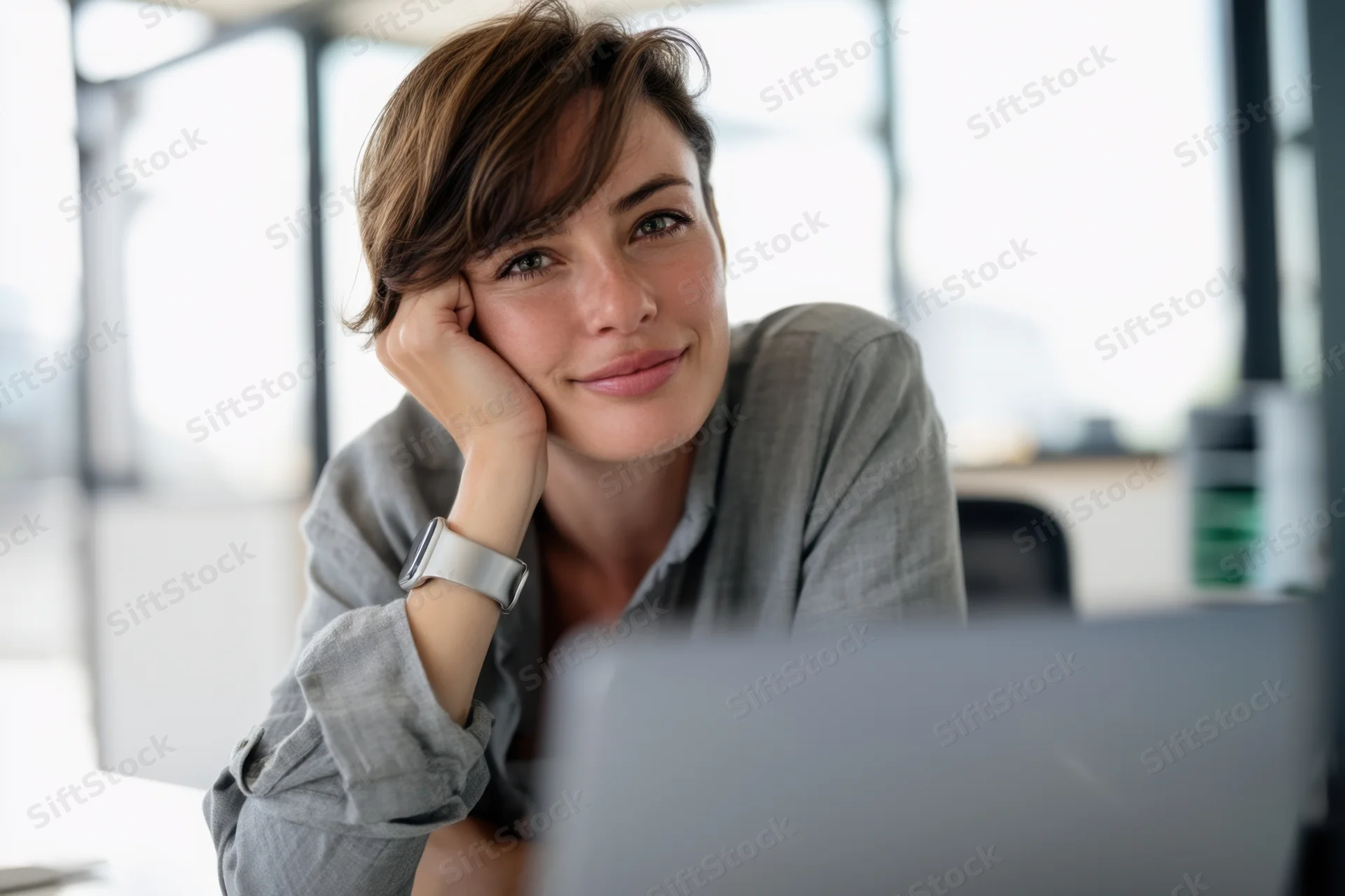 Young woman smiling thoughtfully at desk in office with laptop and smartwatch