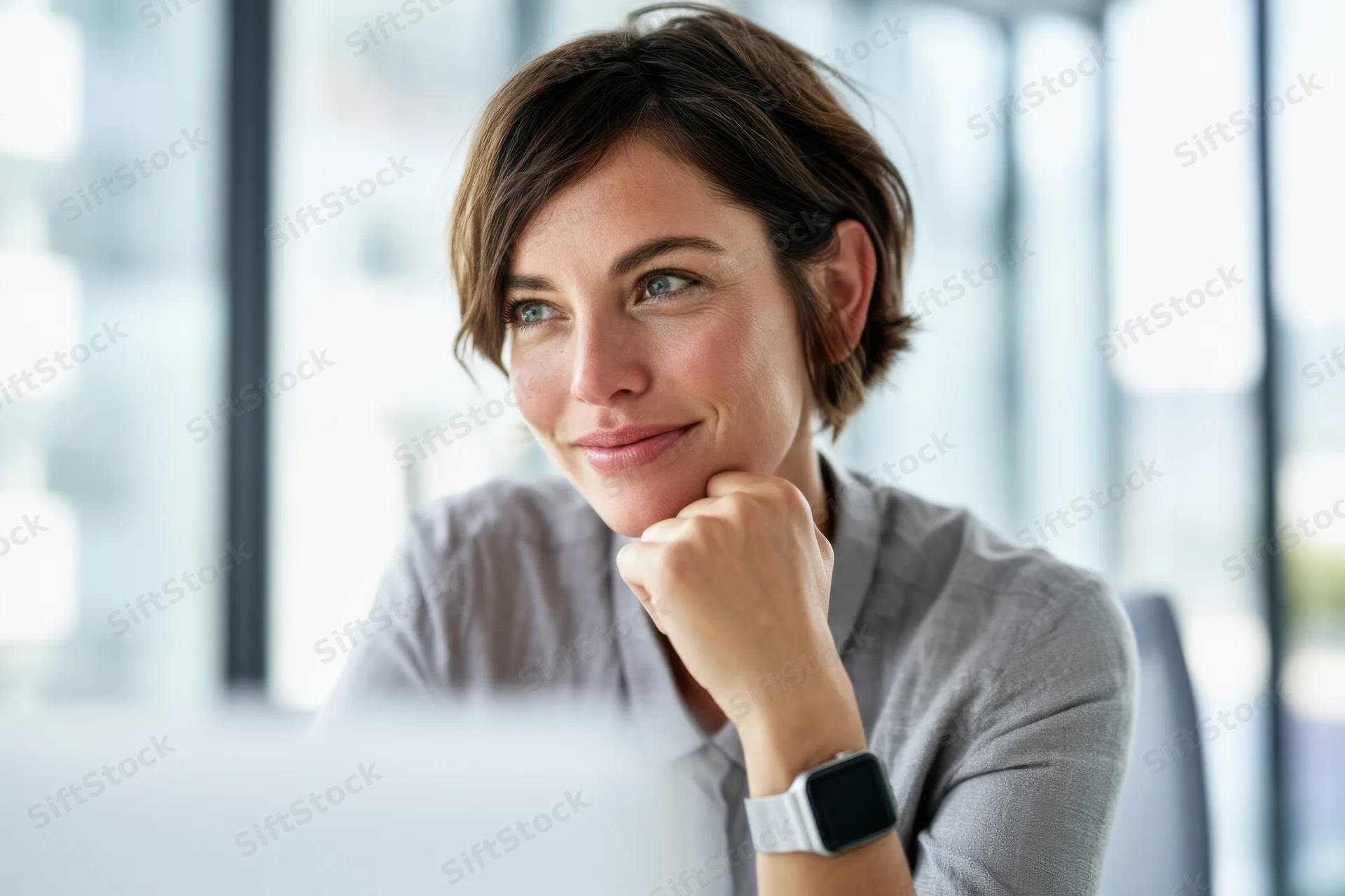 Thoughtful young woman sitting at desk in office with hand on chin, wearing smartwatch and gray shirt, looking off to side