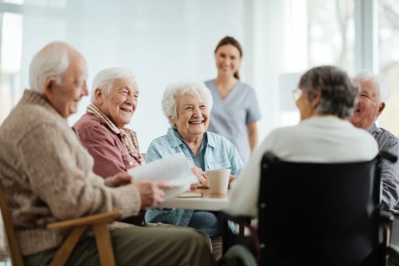 Elderly people smiling and interacting around a table in a bright living room with a caregiver present