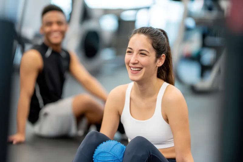 Woman smiling while sitting on floor with foam roller in gym, man in background also exercising