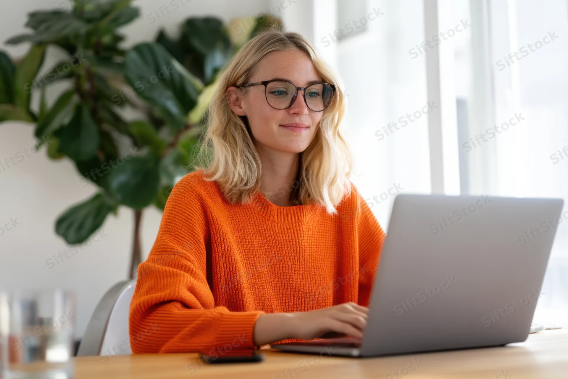 Woman working on laptop at wooden desk in bright home office with plant and window
