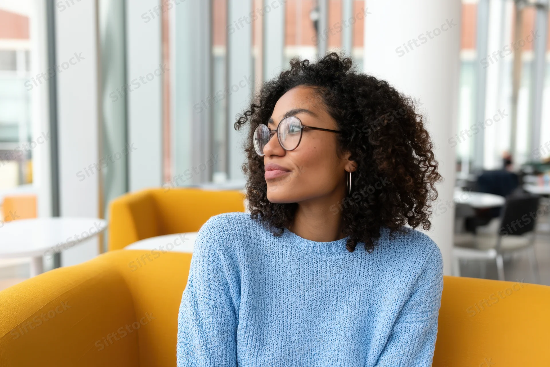Woman with curly hair and round glasses sitting on yellow couch in modern office space smiling