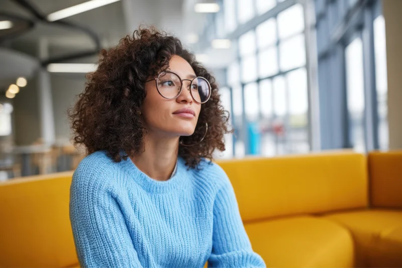 Woman with curly hair and round glasses sitting on yellow couch in modern office interior