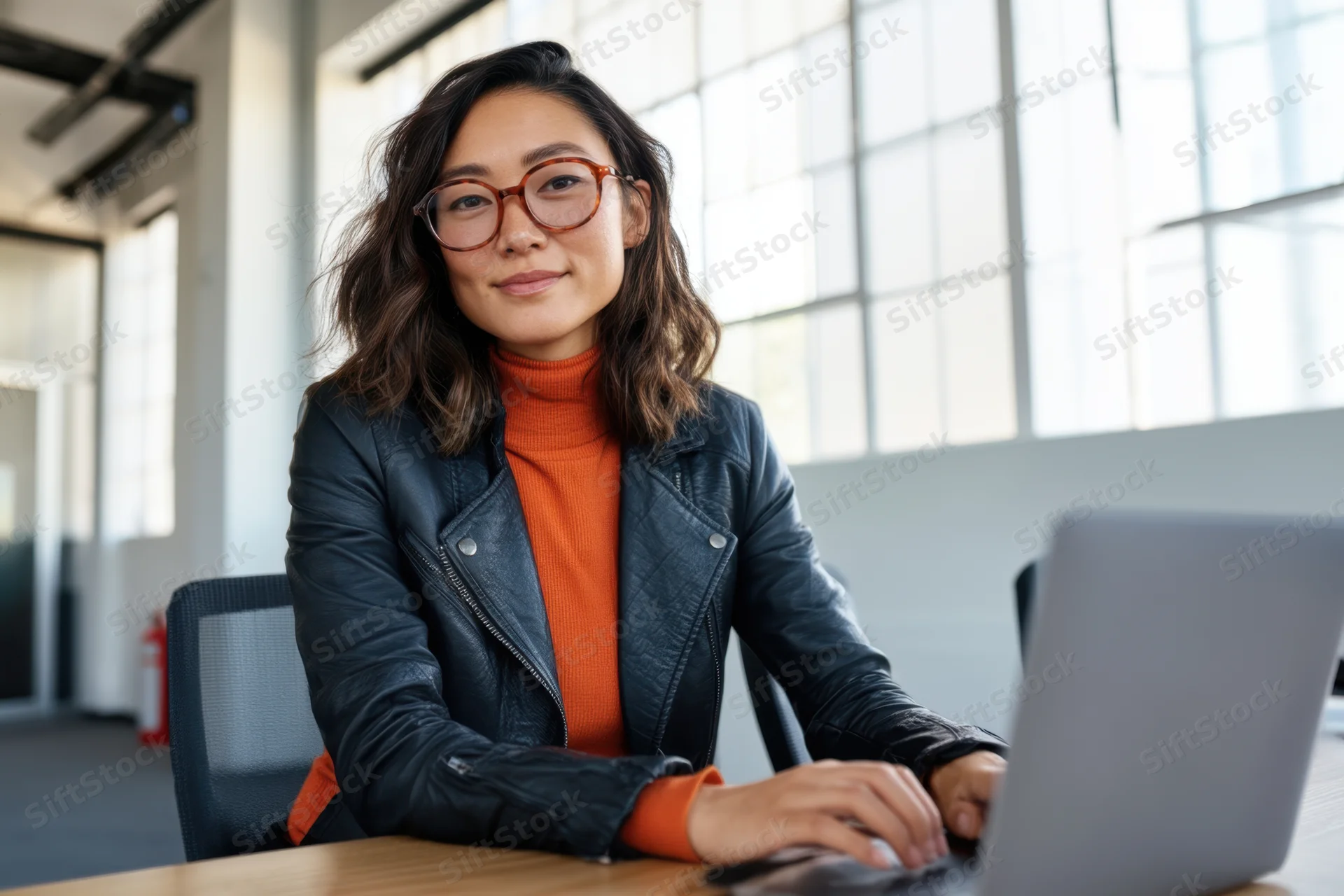 Asian woman in leather jacket and glasses working on laptop at wooden desk in modern office