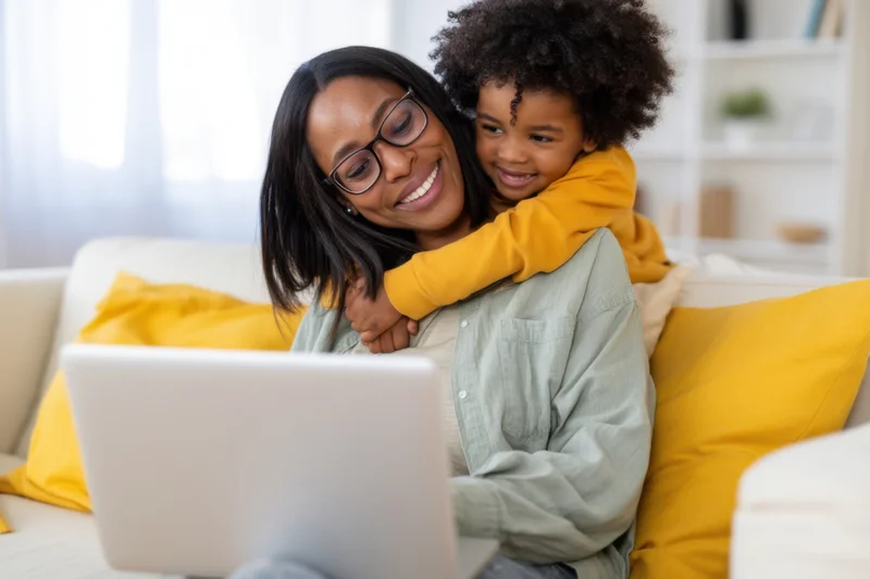 Woman smiling with child hugging her from behind while using laptop on sofa