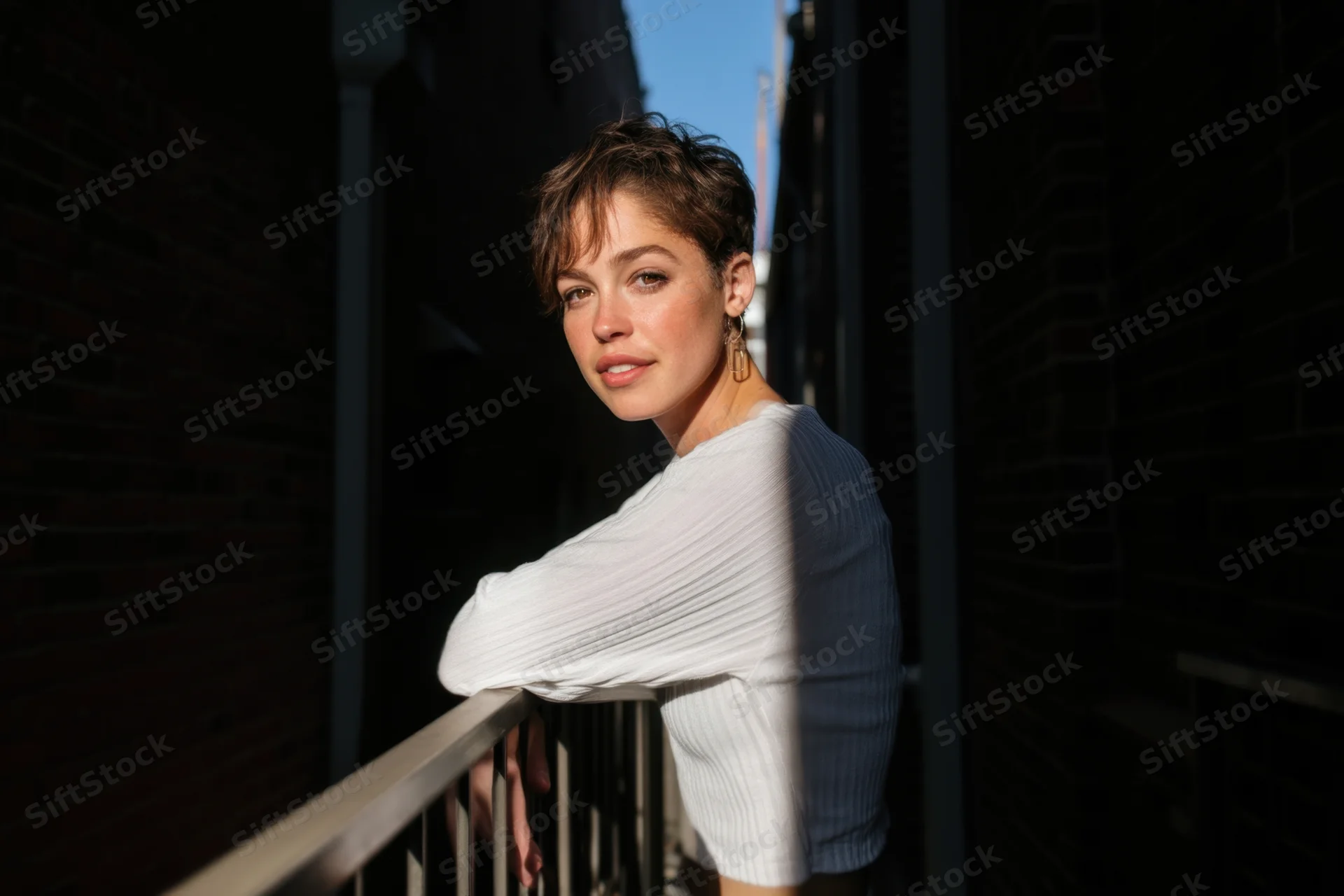Young woman with short hair leaning on balcony railing in urban setting, wearing white ribbed shirt, sunlight and shadow on brick walls